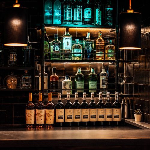Bottiglie di vino allineate sul bancone di ROSSO, davanti alle mensole illuminate di gin, whisky e amari