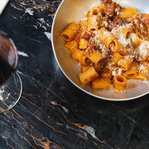 Rigatoni all'amatriciana con pecorino serviti in padella, accanto a un calice di vino rosso